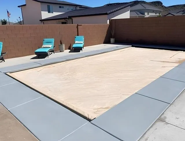 Pool with tan cover, surrounded by gray concrete, lounge chairs nearby. Brown fence and house in background.