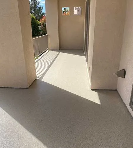 A light-colored balcony with a textured floor, flanked by beige pillars and walls. Sunlight illuminates part of the floor.